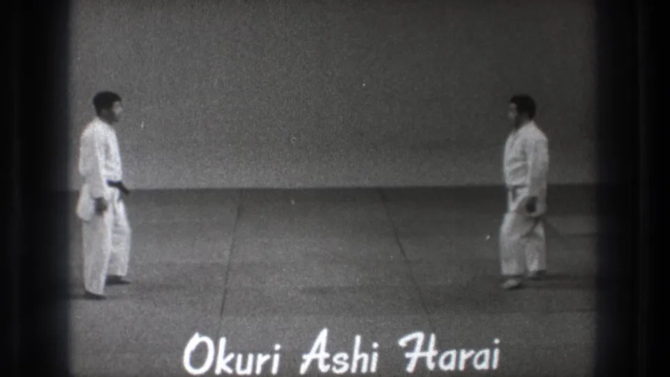 In 1971 Tokyo footage, two adult men in white martial arts gi stand apart on a plain dojo mat as handwritten text reads Okuri Ashi Harai.