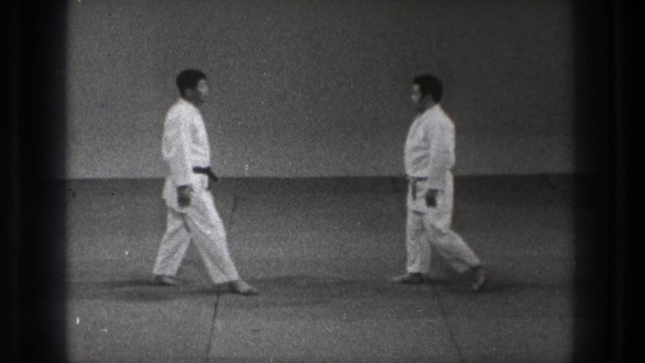 Two adult martial arts practitioners in white gi approach each other in a sparse Tokyo dojo setting on grainy 8mm footage.