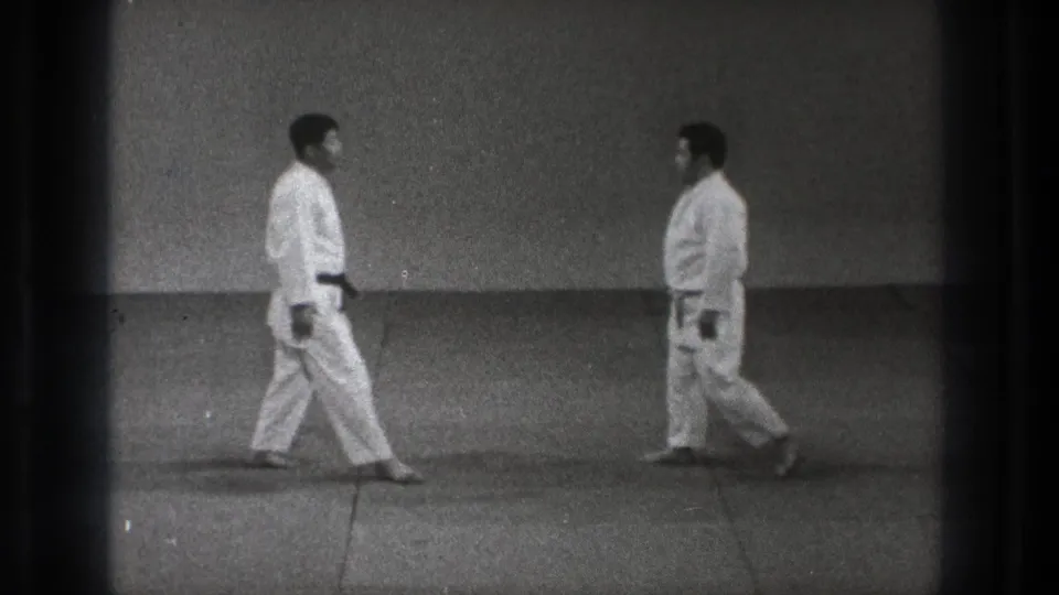 Two adult martial arts practitioners in white gi approach each other in a sparse Tokyo dojo setting on grainy 8mm footage.