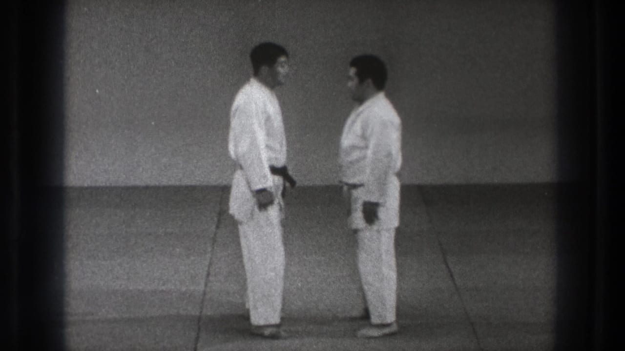 In the dojo sequence, two adult men in white gi stand face to face at close distance on a plain matted floor.