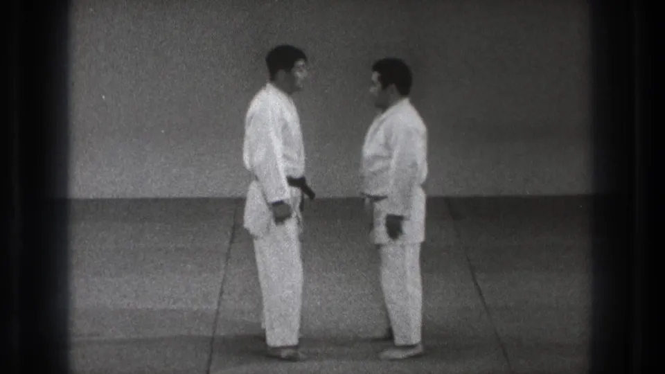 In the dojo sequence, two adult men in white gi stand face to face at close distance on a plain matted floor.