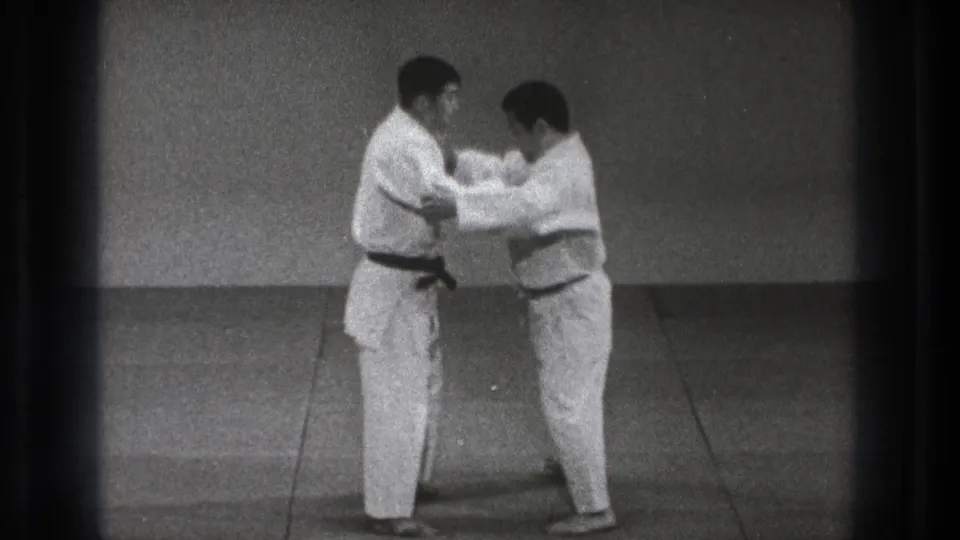 Two adult men in gi begin contact, with one practitioner holding or adjusting the other's uniform during the 1971 Tokyo dojo footage.
