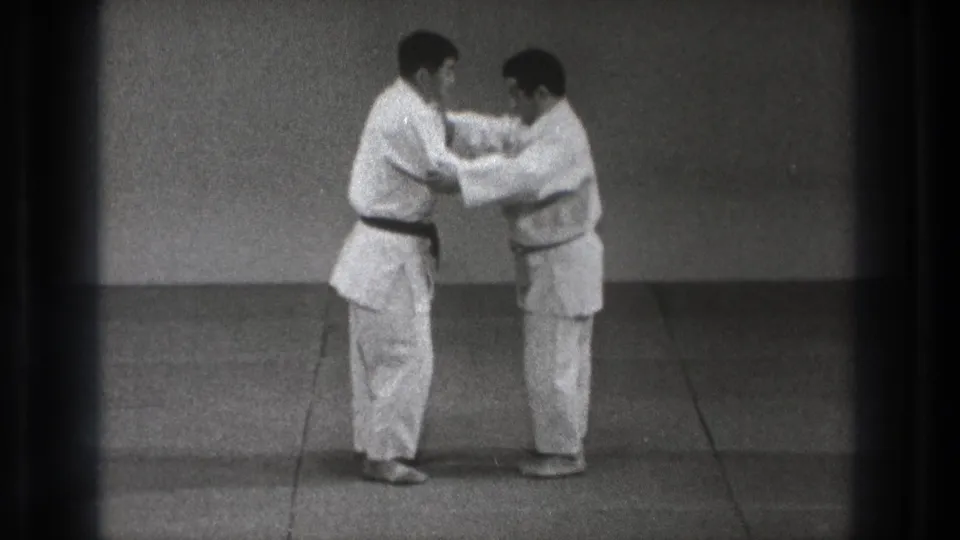 In supported 1971 Tokyo footage, two adult martial artists in white gi and dark belts begin a close grappling drill on a plain mat.