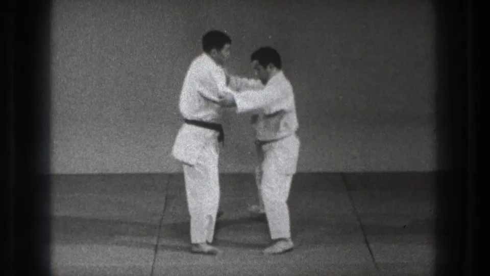 The two adult practitioners continue a controlled karate or judo-style grappling movement in the plain indoor training space.