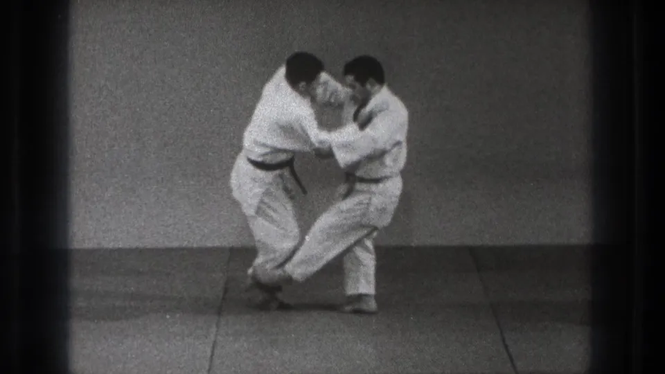 The sequence reaches a controlled off-balance position as one practitioner lifts a leg while both maintain close grips on the mat.