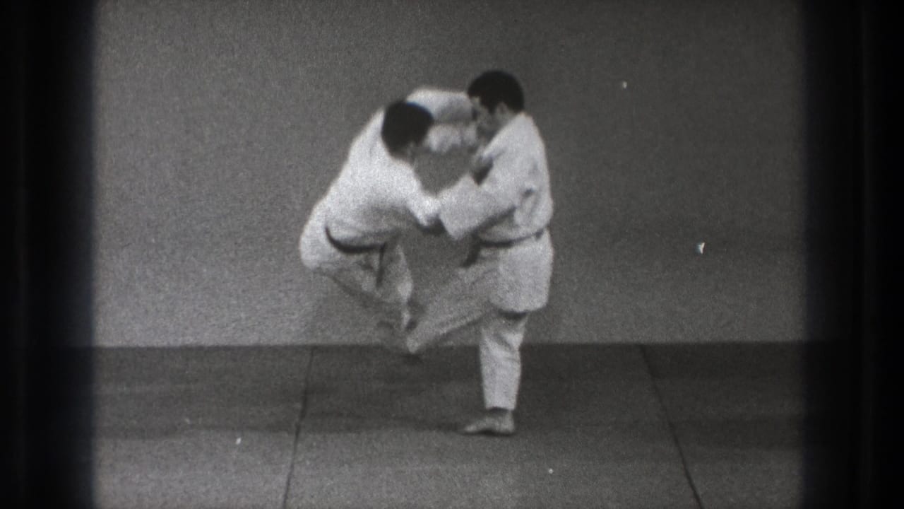 In a Tokyo 1971 martial arts practice setting, two adult men in white gi grip each other as one starts a throw on the mat.