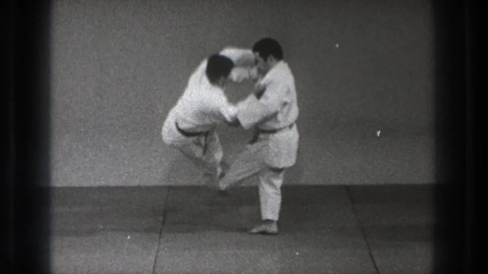 In a Tokyo 1971 martial arts practice setting, two adult men in white gi grip each other as one starts a throw on the mat.