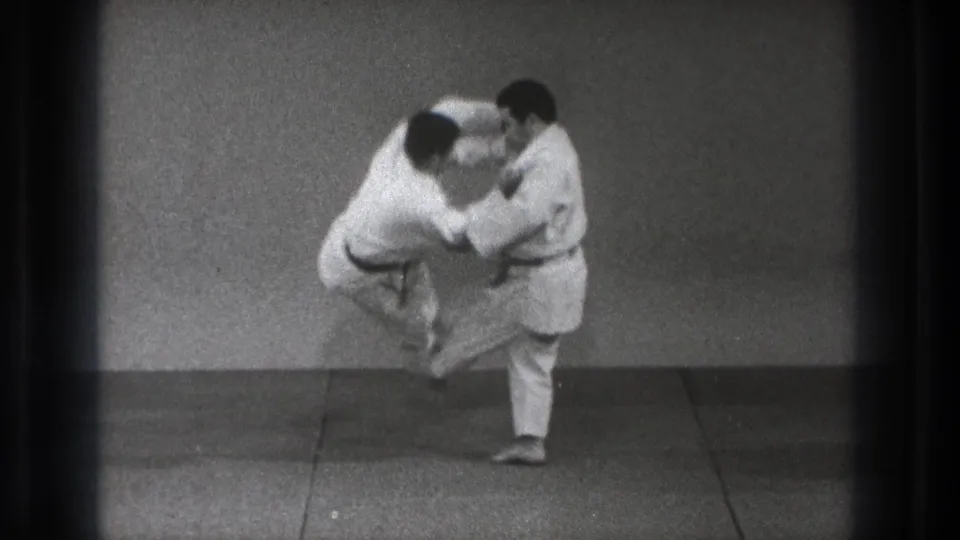 One man in a white gi lifts his practice partner off the mat during a martial arts throw in the Tokyo 1971 footage.
