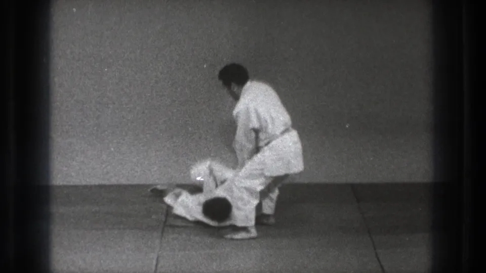 After the takedown, one man in a white gi stands while his partner lies on the mat in the Tokyo 1971 martial arts practice clip.
