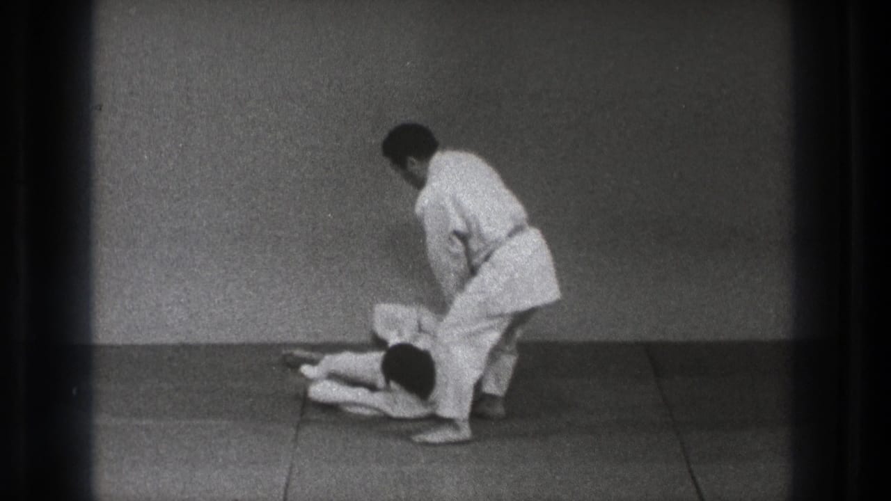 1971 Tokyo 8mm frame showing two adult men in white judogi practicing a floor control movement on a dojo mat.