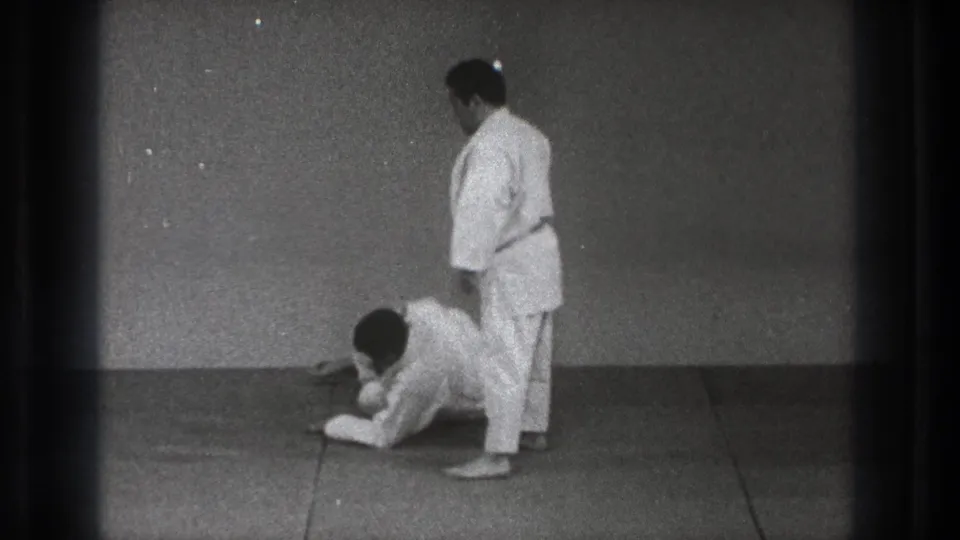 In the Tokyo dojo scene, one adult trainee remains low on the mat while his partner stands over him after a practice movement.