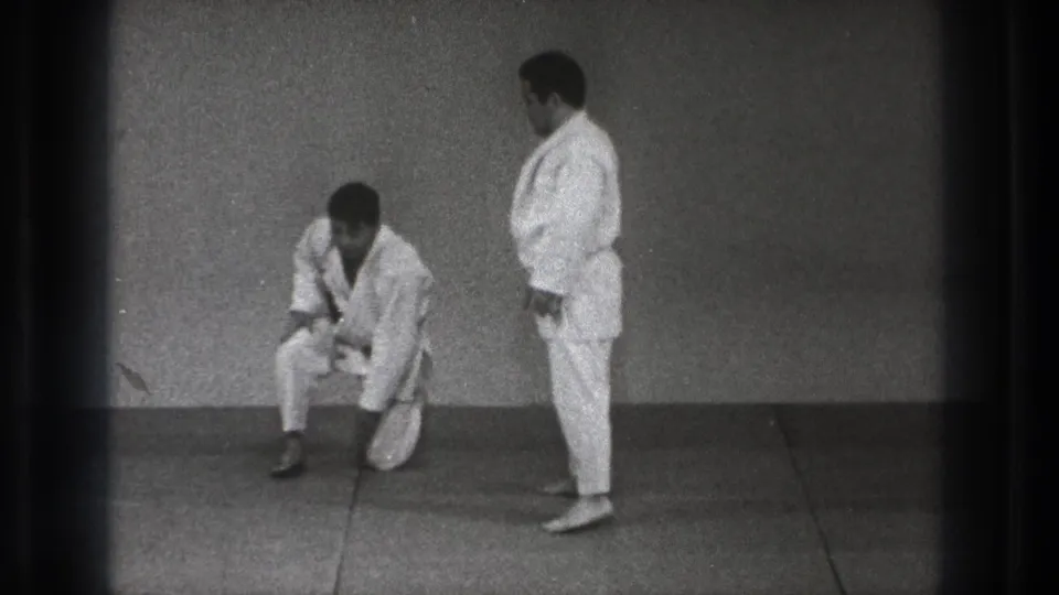 1971 Tokyo judo practice frame with one man kneeling or rising while another stands beside him in a simple dojo.