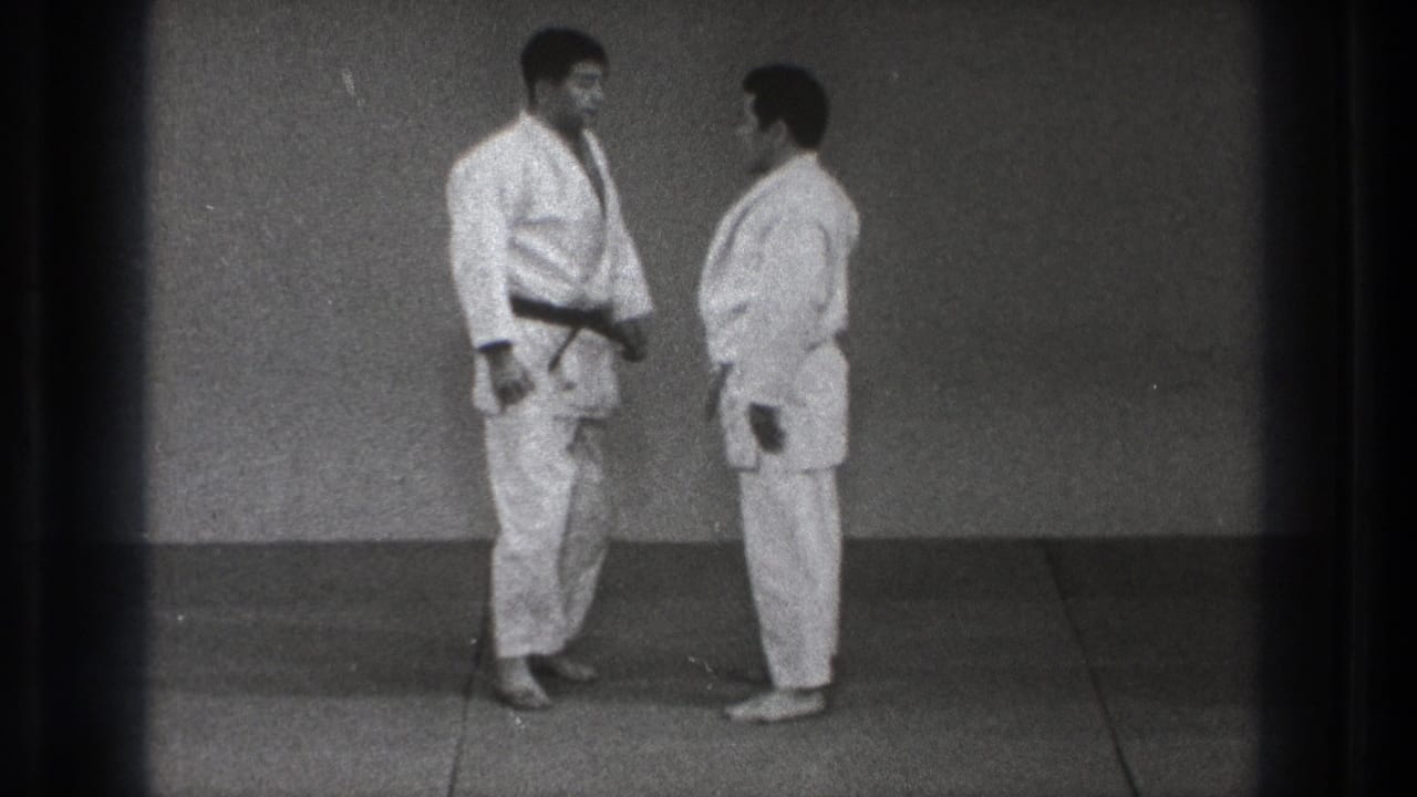 In Tokyo, 1971, two adult men in white karate gi face each other on a plain dojo mat, one wearing a dark belt.