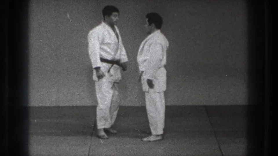 In Tokyo, 1971, two adult men in white karate gi face each other on a plain dojo mat, one wearing a dark belt.