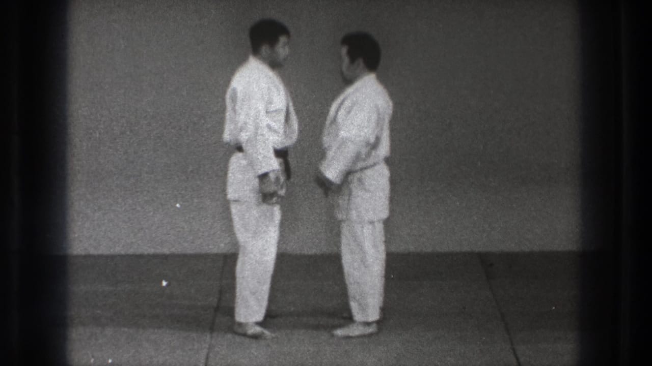 The two adult practitioners remain close on the dojo mat, shown in profile with plain wall and film gate shadow visible.