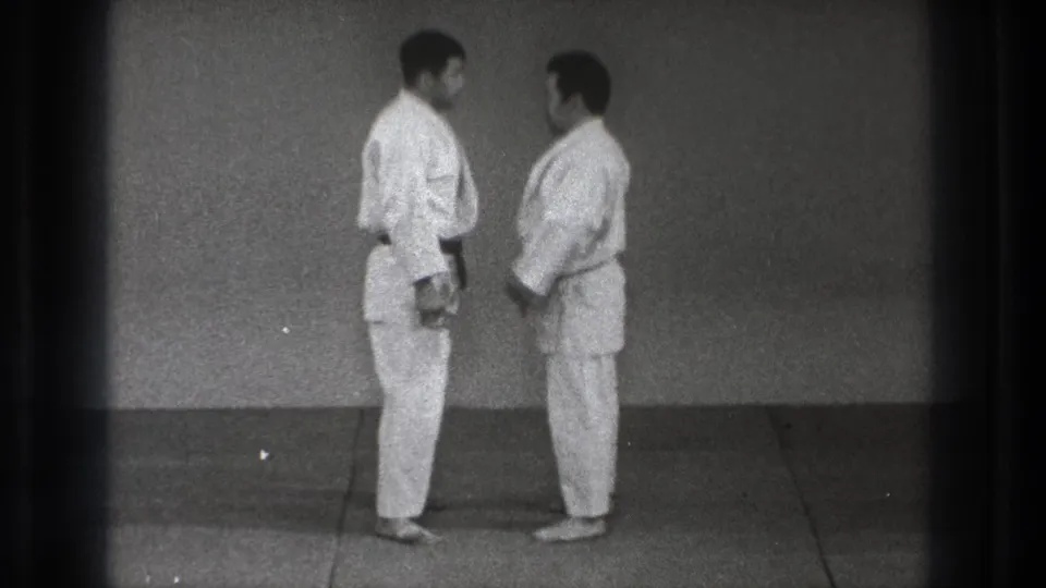 The two adult practitioners remain close on the dojo mat, shown in profile with plain wall and film gate shadow visible.