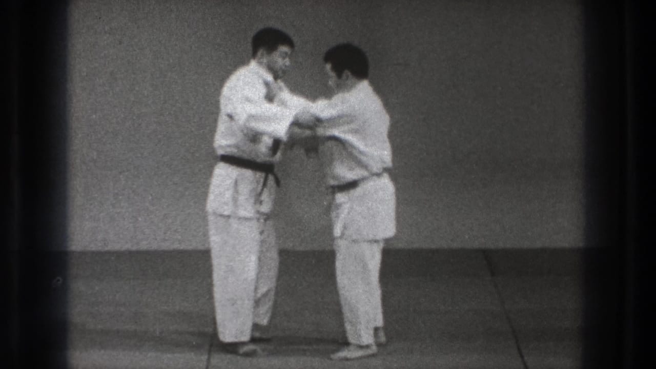 In the Tokyo dojo footage, two adult men in gi take a close grip, with arms raised as a hold or throw setup begins.