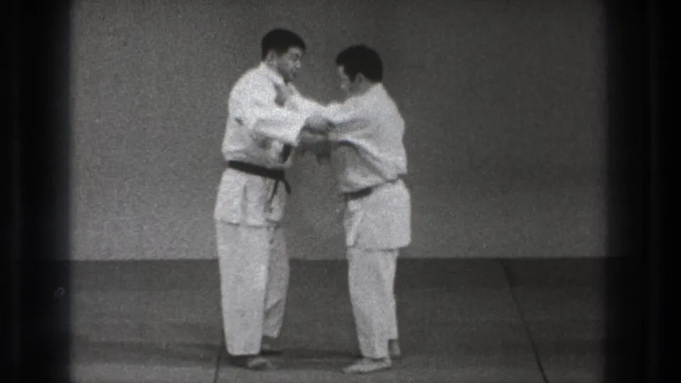 In the Tokyo dojo footage, two adult men in gi take a close grip, with arms raised as a hold or throw setup begins.