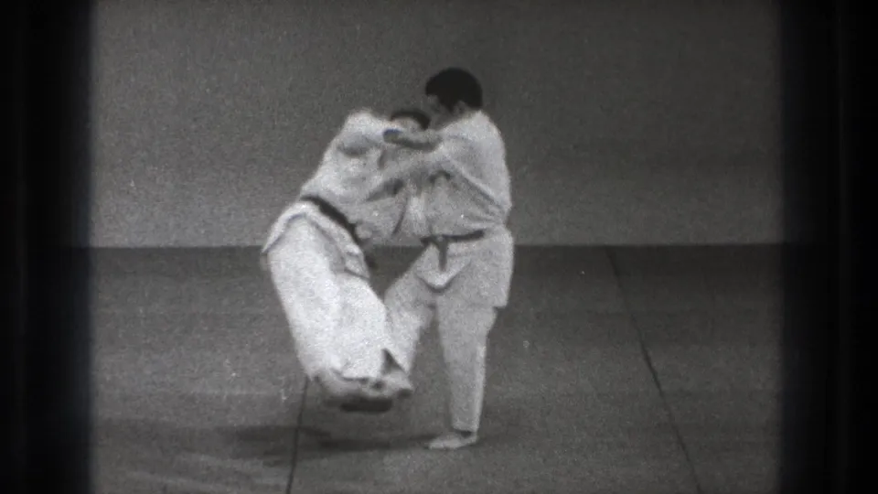 The dark-belt practitioner lifts and turns his partner during a controlled judo throw in the 1971 Tokyo home-movie scene.