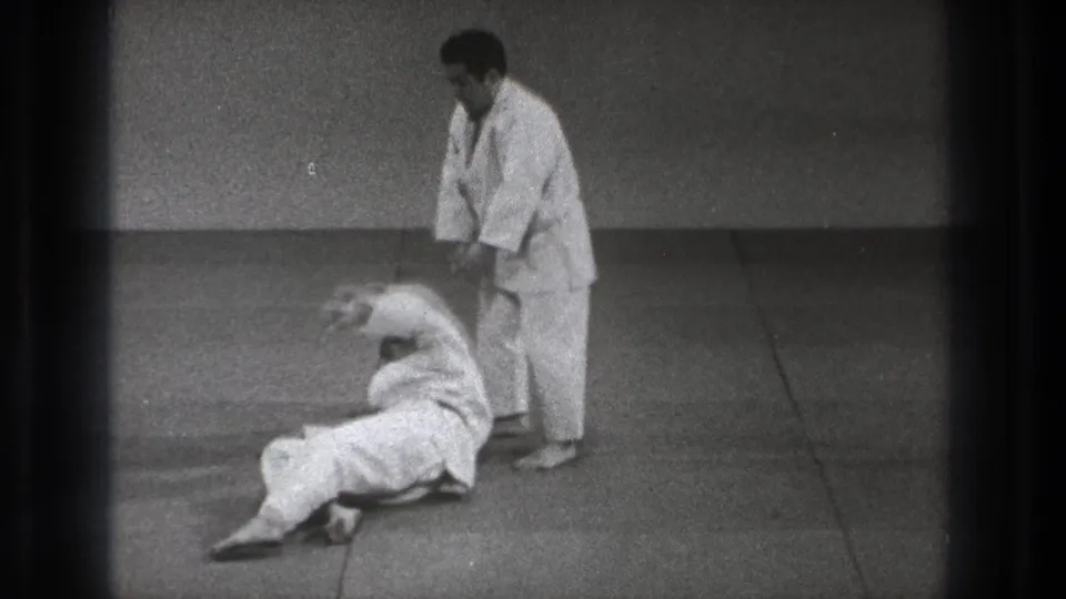 In Tokyo 1971 home-movie footage, one man in a white uniform stands as another falls or rolls on a dojo mat.