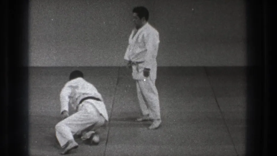 A man in a white martial arts uniform gets up from the mat while his partner stands beside him in the dojo.