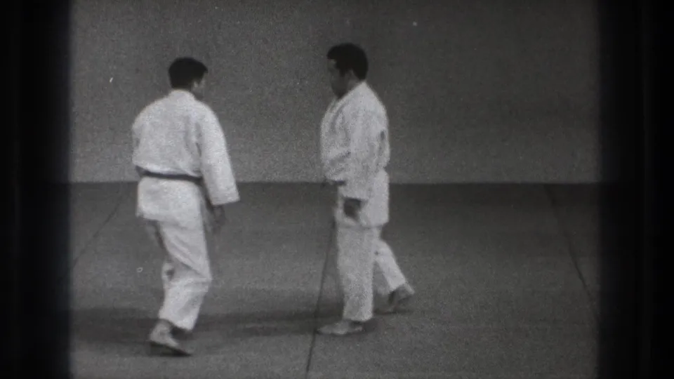Two adult men in white martial arts uniforms reset their stance on a dojo mat in Tokyo 1971 footage.