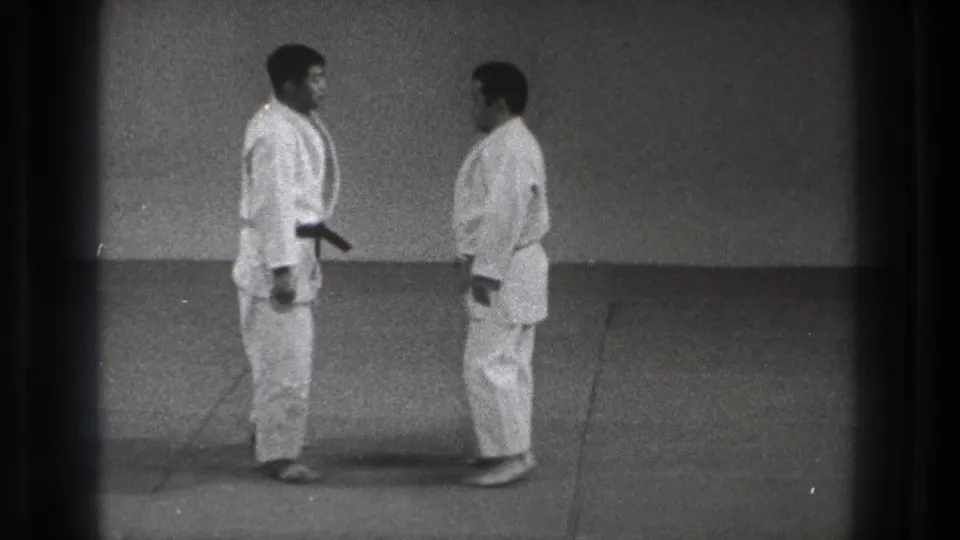 The two men stand face to face in a simple dojo, their white uniforms and mat floor visible in the 1971 Tokyo clip.