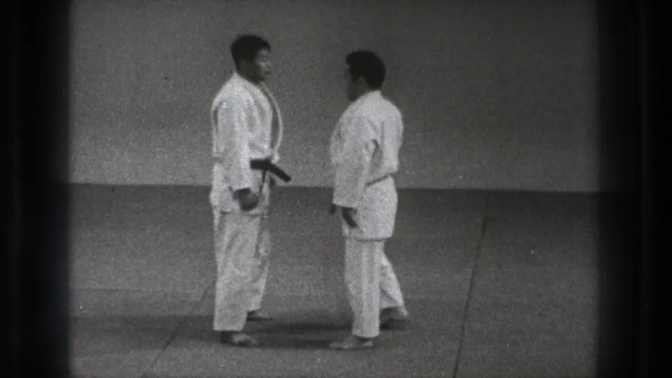 In Tokyo 1971 footage, two adult men in white martial arts uniforms and black belts stand facing each other on a plain training mat.