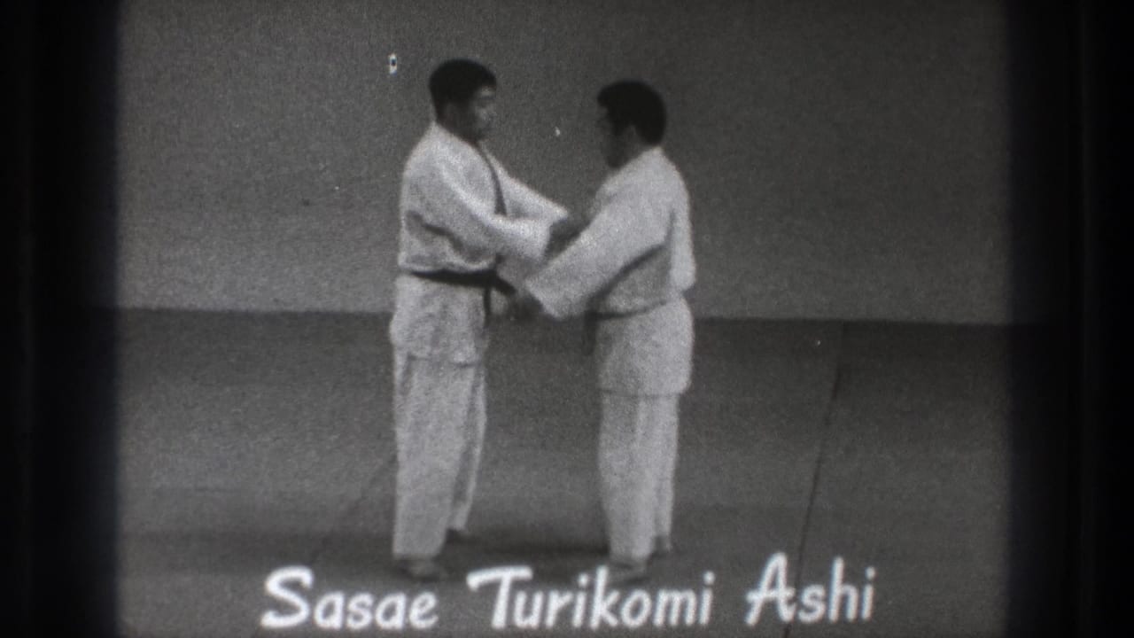 The two men make contact during the demonstration, with the title Sasae Turikomi Ashi visible along the lower edge of the frame.