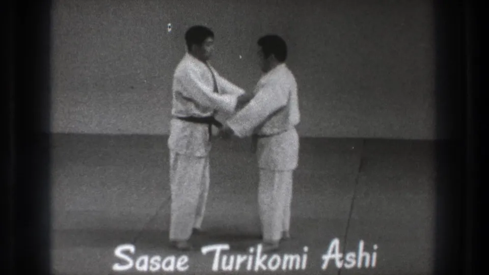 The two men make contact during the demonstration, with the title Sasae Turikomi Ashi visible along the lower edge of the frame.