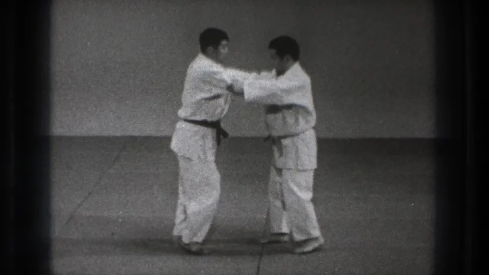 The men hold sleeve and lapel positions while facing each other in the black-and-white 8mm martial arts demonstration.