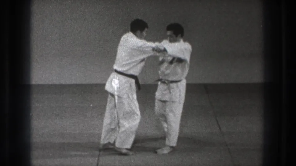 A participant steps across while holding the other man's gi, suggesting the start of the named throwing or foot-blocking technique.