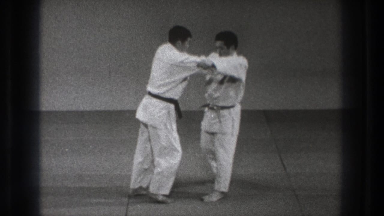 Two men in white gi face each other on mats in a plain training room.