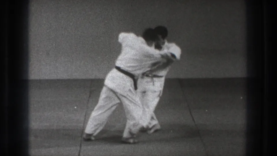 The black-belt practitioner appears to rotate into a throw as his partner bends forward on the mat.