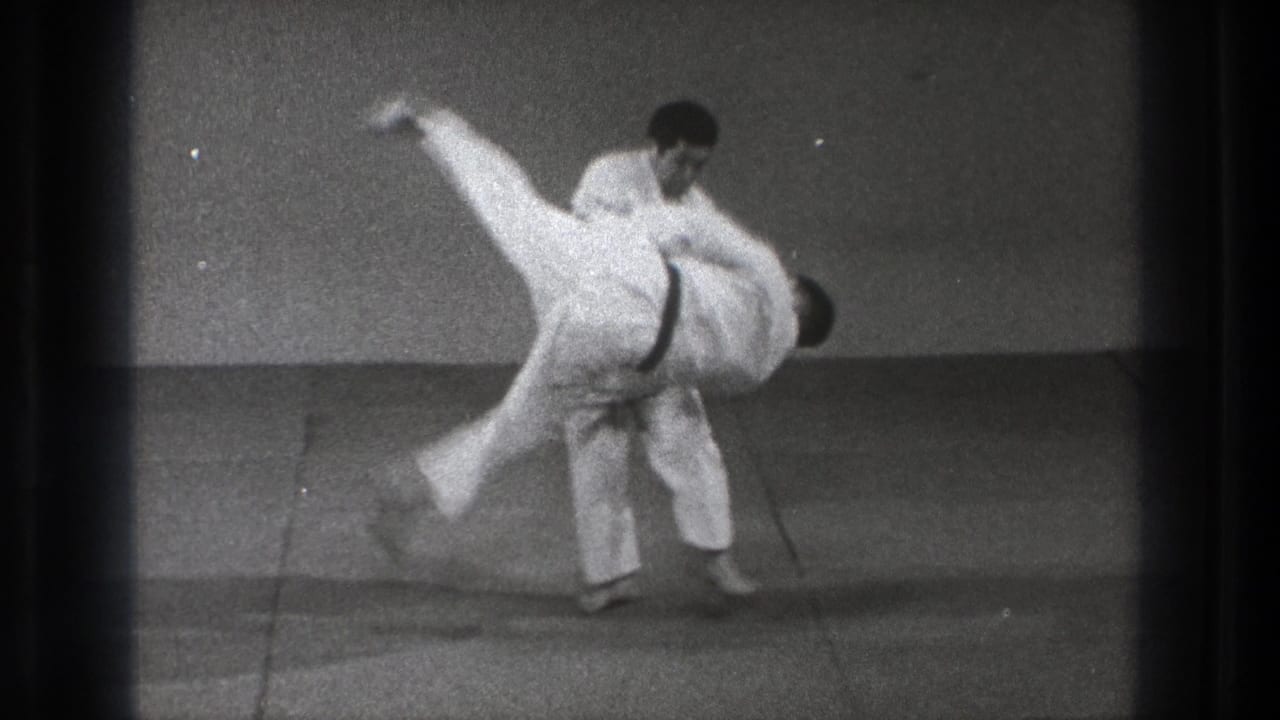 1971 Tokyo 8mm frame showing one adult in a white gi lifting another during a controlled martial arts takedown.