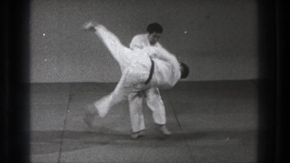 1971 Tokyo 8mm frame showing one adult in a white gi lifting another during a controlled martial arts takedown.