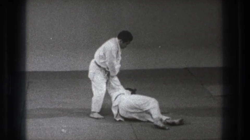 Black-and-white 8mm frame from Tokyo showing the end of a martial arts takedown with both adults controlled on the mat.