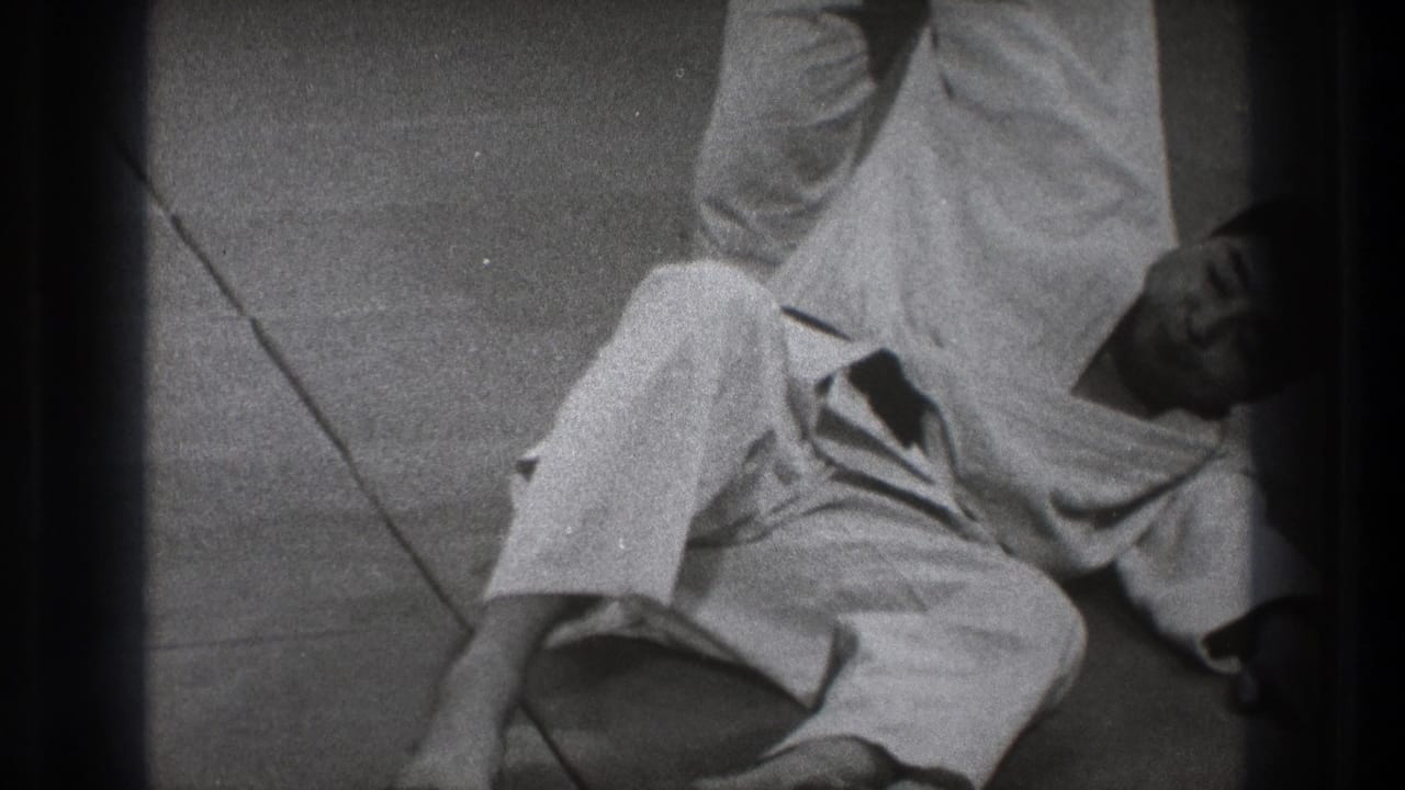 Black-and-white 8mm frame from Tokyo showing a gi-clad karate practitioner low on the mat during a demonstration sequence.