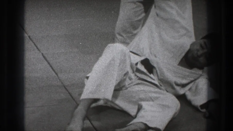 Black-and-white 8mm frame from Tokyo showing a gi-clad karate practitioner low on the mat during a demonstration sequence.
