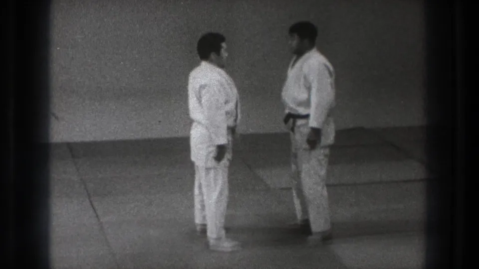 Two adult karate practitioners stand facing each other on a mat in the metadata-supported Tokyo 1971 home-movie footage.