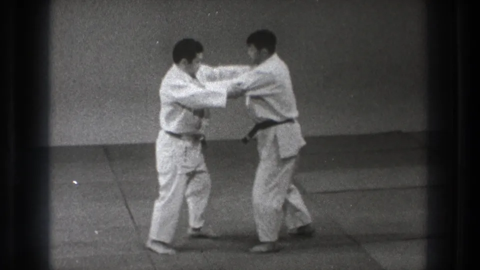 In a Tokyo 1971 8mm frame, two men in white martial arts uniforms stand on a mat while beginning a grip-based defense movement.