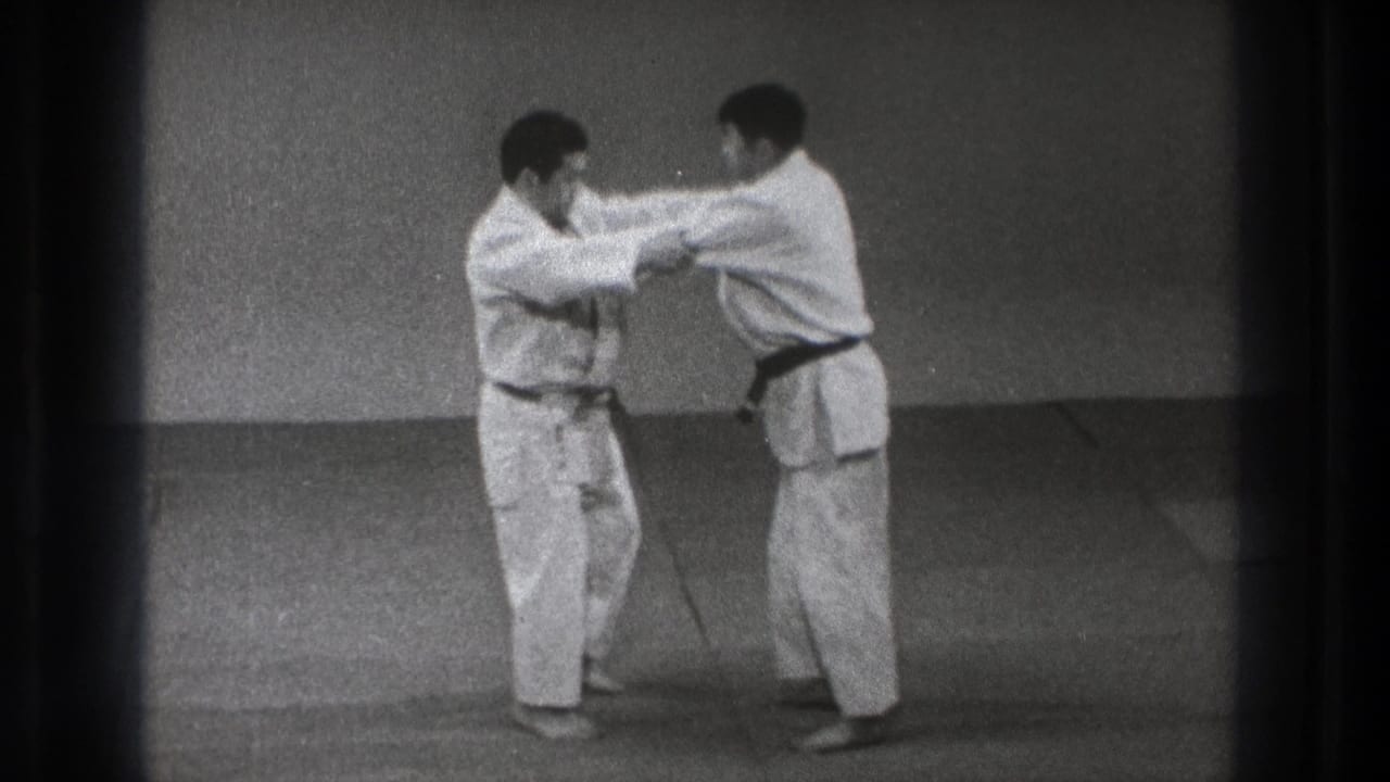 The practitioners remain upright in the dojo setting, maintaining sleeve and arm contact during a controlled defense sequence.