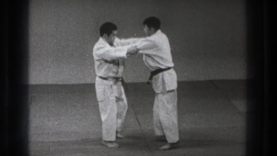 The practitioners remain upright in the dojo setting, maintaining sleeve and arm contact during a controlled defense sequence.