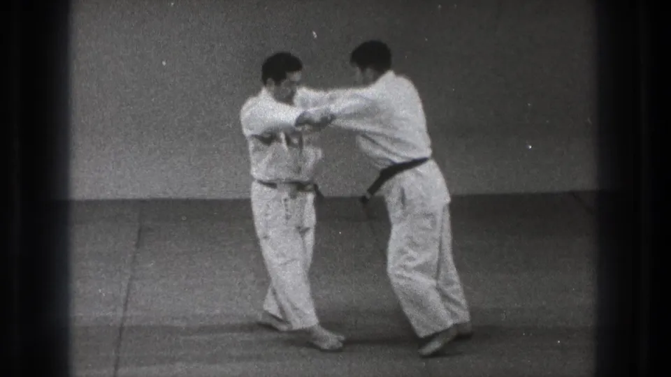During the 8mm training scene, one practitioner extends his arms while the other receives or redirects the movement on the dojo mat.