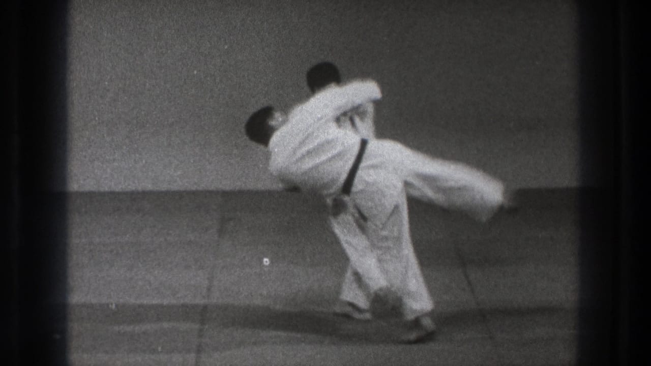 1971 Tokyo 8mm frame showing two adult men in white gi as one begins lifting the other during an indoor martial arts throw.