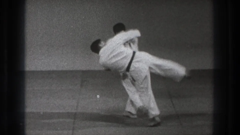 1971 Tokyo 8mm frame showing two adult men in white gi as one begins lifting the other during an indoor martial arts throw.
