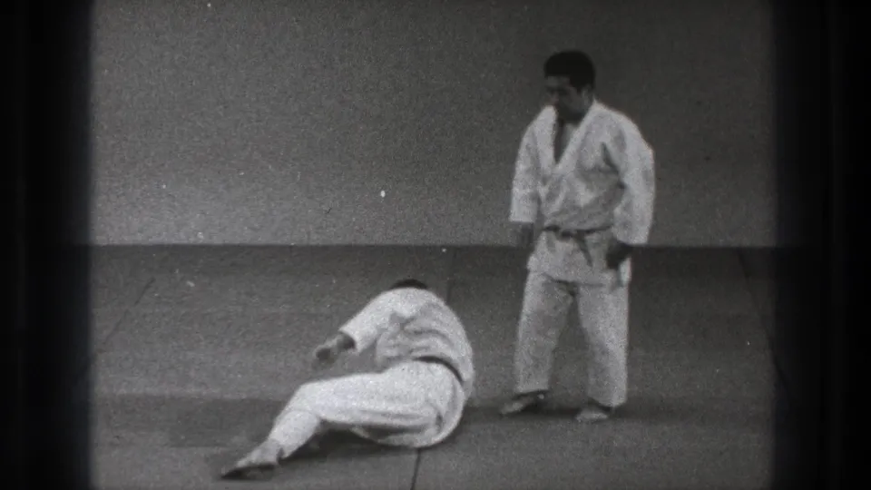 In 1971 Tokyo footage, two adult martial artists in white gi are on dojo mats as one sits low on the floor and the other stands nearby.