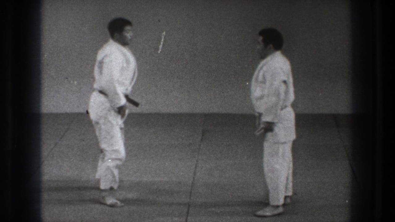 Two adult martial artists in white gi face each other in a plain Tokyo dojo setting, appearing to reset for the next practice move.