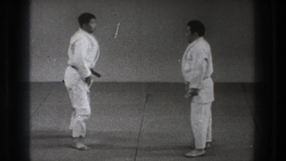 Two adult martial artists in white gi face each other in a plain Tokyo dojo setting, appearing to reset for the next practice move.