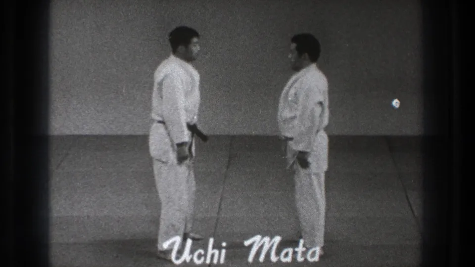In 1971 Tokyo footage, two adult martial arts practitioners in white gi stand facing each other on a plain indoor mat with 'Uchi Mata' on screen.